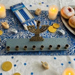 May include: A Hanukkah arrangement with a bronze menorah, lit candles, and dreidels. Chocolate coins and a box of blue and white candles are scattered around. Sufganiyot, or jam doughnuts, are on a silver platter.