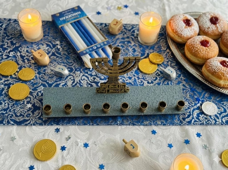 May include: A Hanukkah arrangement with a bronze menorah, lit candles, and dreidels. Chocolate coins and a box of blue and white candles are scattered around. Sufganiyot, or jam doughnuts, are on a silver platter.