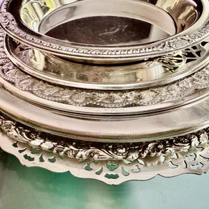May include: A stack of four round silver-toned metal plates and trays. The top piece is a deep bowl, followed by three flat plates of varying sizes, each featuring intricate embossed floral and scrollwork borders with scalloped edges.