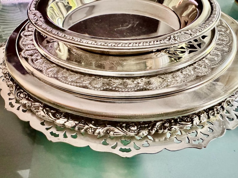 May include: A stack of four round silver-toned metal plates and trays. The top piece is a deep bowl, followed by three flat plates of varying sizes, each featuring intricate embossed floral and scrollwork borders with scalloped edges.
