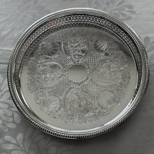 May include: A round silver-toned metal tray featuring an intricate embossed floral and scrollwork pattern across the center. The tray has a raised, openwork lattice border and a twisted rope-style edge detail around the entire circumference.