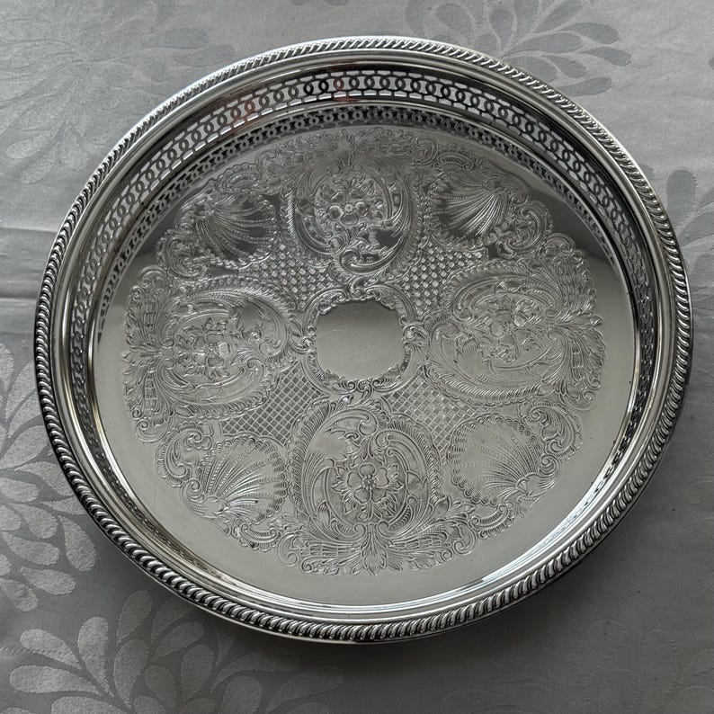 May include: A round silver-toned metal tray featuring an intricate embossed floral and scrollwork pattern across the center. The tray has a raised, openwork lattice border and a twisted rope-style edge detail around the entire circumference.