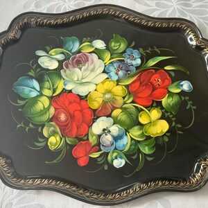 May include: A black serving tray with a scalloped edge and a gold trim. The tray is decorated with a vibrant floral arrangement featuring red, white, yellow, and blue flowers with green leaves.