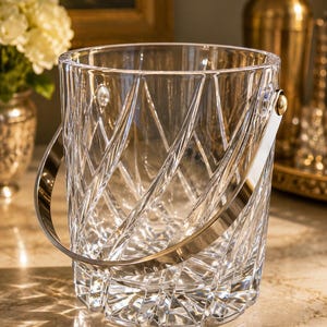 May include: A clear glass ice bucket featuring a deep, crisscross geometric cut pattern across its exterior. The bucket includes a polished silver-tone metal swing handle attached to the sides with circular hardware, creating a functional design.