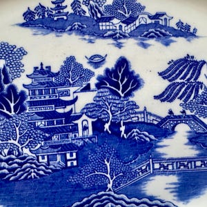 K&ouml;nnte beinhalten: Blaue und wei&szlig;e Keramikplatte mit einem traditionellen chinesischen Weidenmuster. Das Design zeigt eine Pagode, eine Br&uuml;cke, B&auml;ume und einen Fluss.