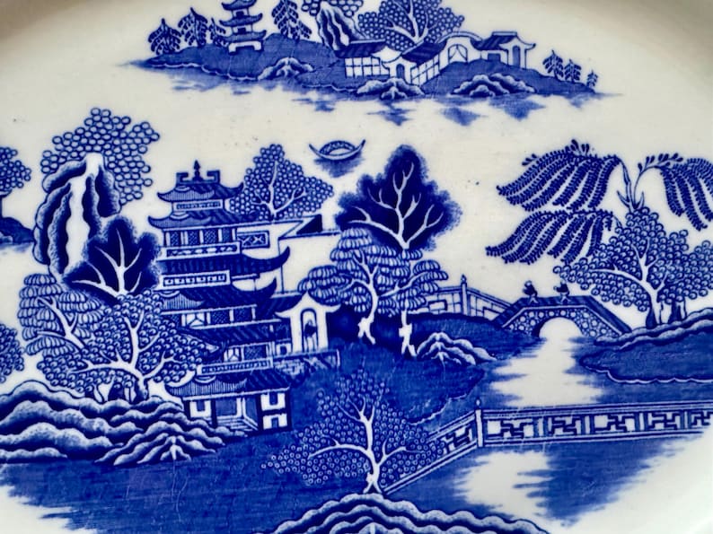 K&ouml;nnte beinhalten: Blaue und wei&szlig;e Keramikplatte mit einem traditionellen chinesischen Weidenmuster. Das Design zeigt eine Pagode, eine Br&uuml;cke, B&auml;ume und einen Fluss.