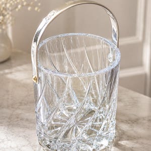 May include: A clear glass ice bucket featuring a deep, swirling geometric cut pattern across the exterior. The cylindrical vessel includes a polished silver-toned metal handle attached to the sides with small, circular gold-toned hardware accents.