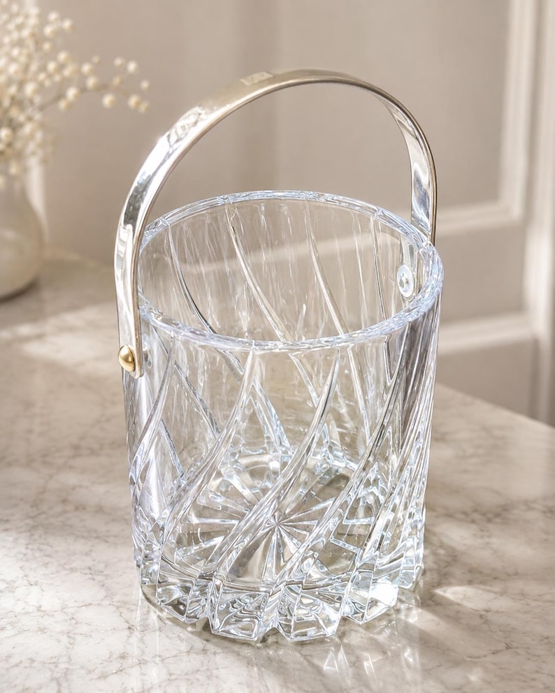 May include: A clear glass ice bucket featuring a deep, swirling geometric cut pattern across the exterior. The cylindrical vessel includes a polished silver-toned metal handle attached to the sides with small, circular gold-toned hardware accents.