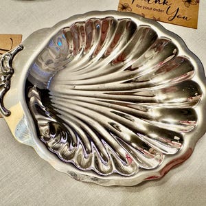 K&ouml;nnte beinhalten: Eine silberfarbene Metallschale in Form einer Jakobsmuschel mit einer polierten, reflektierenden Oberfl&auml;che. Die Schale weist tiefe, strahlenf&ouml;rmig verlaufende Rillen und ein verziertes Griffdetail an einer Seite auf, das die nat&uuml;rliche Form nachahmt.