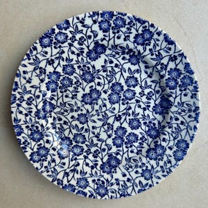 May include: A round ceramic plate featuring a dense, repeating pattern of small blue flowers and thin, winding stems against a white background. The floral design covers the entire surface of the plate, extending from the center to the rim.