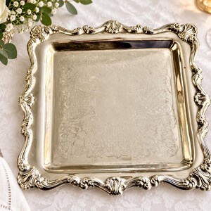 Puede incluir: Una bandeja de servicio cuadrada de tono plateado con un borde elevado y ornamentado con intrincados adornos de volutas y motivos de conchas. La superficie central de la bandeja es lisa y pulida, enmarcada por un patr&oacute;n decorativo en relieve.