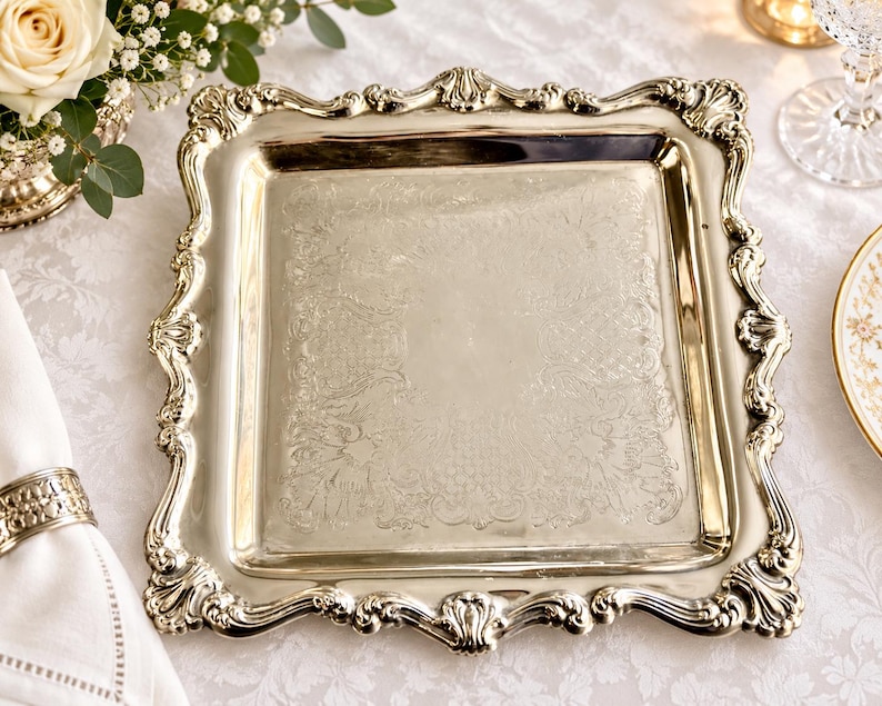 Puede incluir: Una bandeja de servicio cuadrada de tono plateado con un borde elevado y ornamentado con intrincados adornos de volutas y motivos de conchas. La superficie central de la bandeja es lisa y pulida, enmarcada por un patr&oacute;n decorativo en relieve.