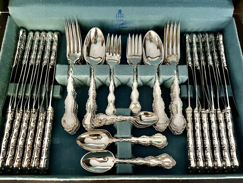 May include: A complete set of ornate silver flatware, including forks, spoons, and knives, arranged in a blue velvet-lined case. The silverware features detailed decorative handles and a polished finish, ideal for formal dining.