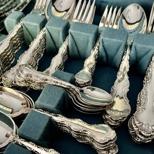 May include: A collection of ornate silver-colored flatware, including spoons, forks, and knives, arranged in a blue velvet-lined case. The silverware features detailed scrollwork and decorative handles. The pieces are neatly organized, suggesting a formal dining set.