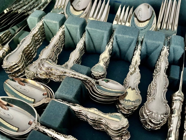 May include: A collection of ornate silver-colored flatware, including spoons, forks, and knives, arranged in a blue velvet-lined case. The silverware features detailed scrollwork and decorative handles. The pieces are neatly organized, suggesting a formal dining set.