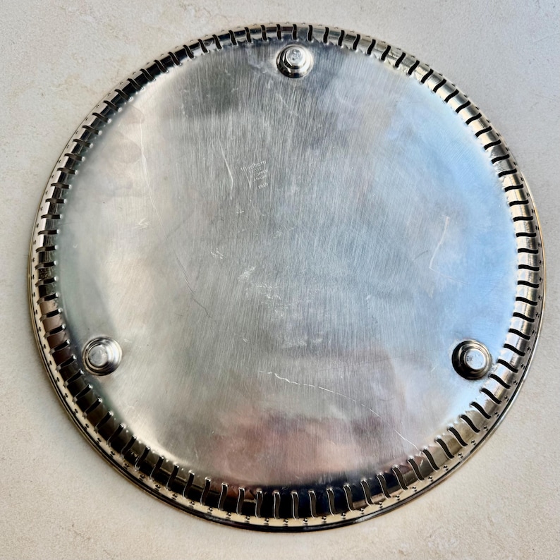 Puede incluir: Una bandeja redonda de metal plateado con un borde decorativo y acanalado. La bandeja tiene tres peque&ntilde;os pies redondos y una superficie lisa y reflectante. El borde presenta una serie de hendiduras verticales. Probablemente se utiliza para servir o exhibir.