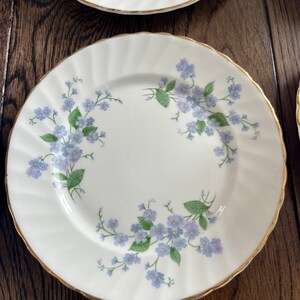 May include: White porcelain plates with a delicate blue floral pattern and gold trim. The plates feature a scalloped edge and are arranged on a dark wood surface. The floral design includes small blue flowers and green leaves.