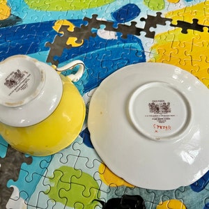Pu&ograve; includere: Un set di tazza e piattino giallo e bianco in porcellana fine. Il lato inferiore della tazza e del piattino presenta un marchio del produttore Paragon stampato in nero con il testo H.M. THE QUEEN & H.M. QUEEN MARY FINE BONE CHINA ENGLAND REG'D.
