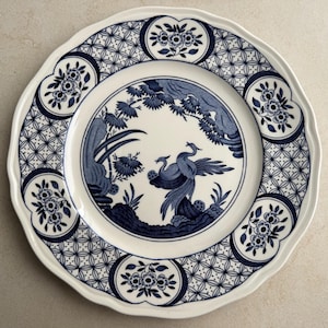 Puede incluir: Un plato redondo de cer&aacute;mica con borde festoneado, con base blanca y patrones de transferencia azul oscuro. El centro muestra dos aves entre follaje, mientras que el borde presenta un dise&ntilde;o de celos&iacute;a repetitivo y medallones florales.