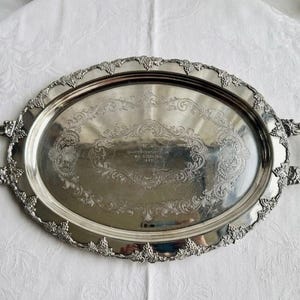 Pu&ograve; includere: Un vassoio da portata ovale in argento con manici e bordo decorati con rilievi di uva e viti. Il centro presenta un'iscrizione incisa che recita Quarter Century Club W.C. Sterling 1948 circondata da intricati motivi floreali e volute.