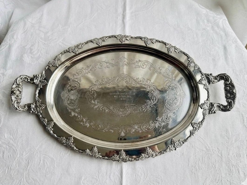 Pu&ograve; includere: Un vassoio da portata ovale in argento con manici e bordo decorati con rilievi di uva e viti. Il centro presenta un'iscrizione incisa che recita Quarter Century Club W.C. Sterling 1948 circondata da intricati motivi floreali e volute.
