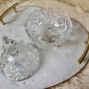 May include: A heart-shaped white marble serving tray with gold-toned metal handles, featuring a clear glass candy dish with a matching lid. The glass dish has a faceted starburst pattern and sits on small feet, displayed with the lid removed.