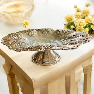 May include: A silver-colored, ornate serving dish with a pedestal base. The dish has a scalloped edge and is decorated with floral patterns. It sits on a light-colored wooden table, with yellow flowers in the background. The dish is approximately 12 inches long.