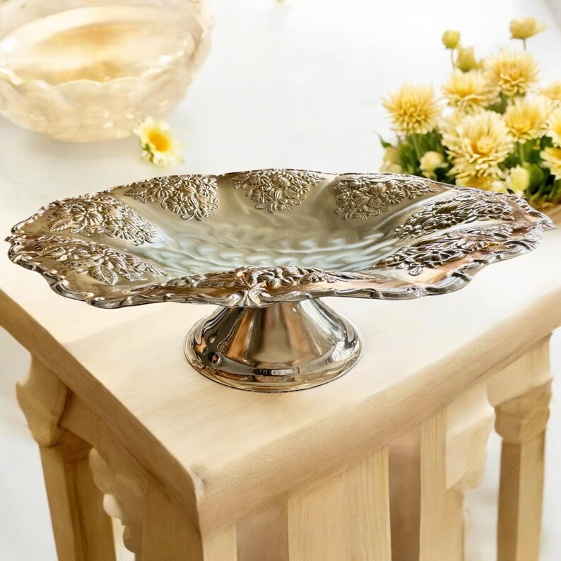 May include: A silver-colored, ornate serving dish with a pedestal base. The dish has a scalloped edge and is decorated with floral patterns. It sits on a light-colored wooden table, with yellow flowers in the background. The dish is approximately 12 inches long.