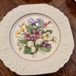 May include: A white ceramic plate with an octagonal shape and a raised, embossed scrollwork border. The center features a colorful botanical illustration of purple, white, and yellow flowers with green leaves, finished with a thin gold rim.