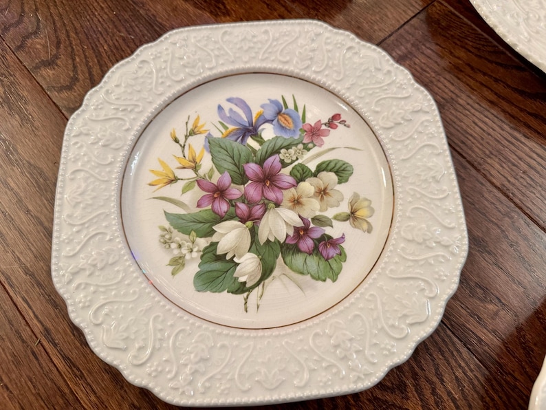 May include: A white ceramic plate with an octagonal shape and a raised, embossed scrollwork border. The center features a colorful botanical illustration of purple, white, and yellow flowers with green leaves, finished with a thin gold rim.
