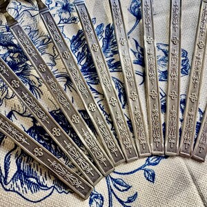 May include: A set of silver-toned metal spoons featuring long, flat handles with an embossed, intricate pattern of scrolling lines and small four-petaled floral motifs. The spoons are arranged in a fan-like formation on a textured surface.