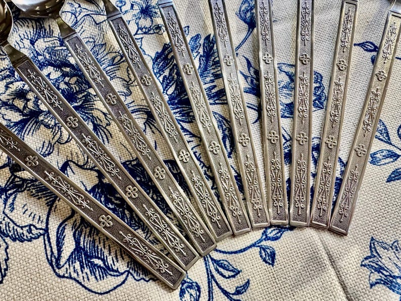 May include: A set of silver-toned metal spoons featuring long, flat handles with an embossed, intricate pattern of scrolling lines and small four-petaled floral motifs. The spoons are arranged in a fan-like formation on a textured surface.
