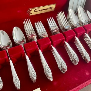 May include: A set of Community silverware, including spoons, forks, and knives, arranged in a red velvet-lined box. The silverware has a floral design on the handles and is displayed neatly. The box has a gold label.