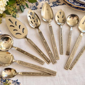 May include: A collection of silver-toned stainless steel flatware and serving utensils featuring ornate, embossed handles with intricate floral and geometric patterns. The set includes various spoons, a cake server, a ladle, and a serving fork.