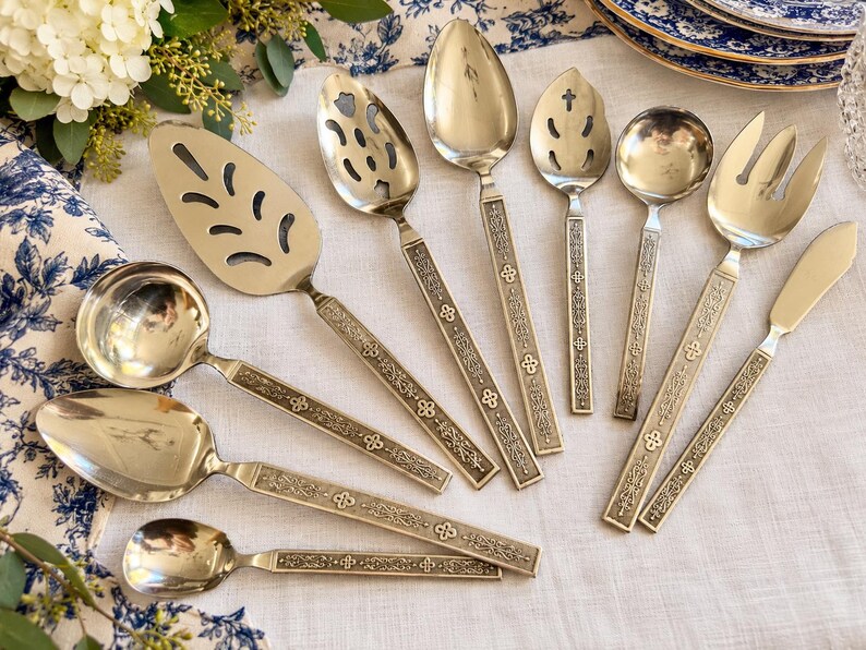 May include: A collection of silver-toned stainless steel flatware and serving utensils featuring ornate, embossed handles with intricate floral and geometric patterns. The set includes various spoons, a cake server, a ladle, and a serving fork.