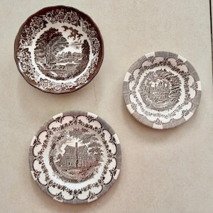 May include: Three ceramic plates featuring brown and white transferware patterns. One bowl displays a rural landscape, while the two smaller plates depict detailed architectural scenes of historic buildings surrounded by intricate floral borders.