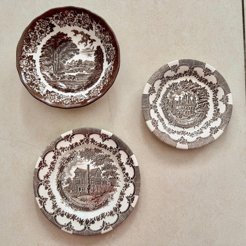 May include: Three ceramic plates featuring brown and white transferware patterns. One bowl displays a rural landscape, while the two smaller plates depict detailed architectural scenes of historic buildings surrounded by intricate floral borders.