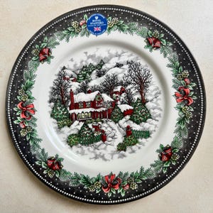 以下が含まれることがあります： ロイヤルスタッフォードのクリスマスプレート。冬のシーンが描かれています。黒いドットの縁に赤いリボンと緑の葉が描かれています。中央には、赤いレンガの家と木々がある雪に覆われた村が描かれています。