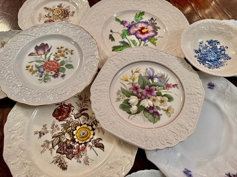 May include: A collection of white ceramic plates and a bowl featuring embossed rims and various colorful floral patterns, including depictions of tulips, roses, irises, and violets, arranged in an overlapping display on a dark wooden surface.