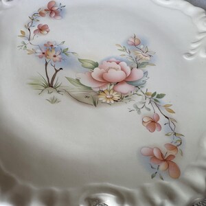 May include: A white ceramic serving platter with a scalloped edge and handles. The platter is decorated with a floral design featuring pink and peach flowers, green leaves, and yellow accents. A decorative piece for serving food.