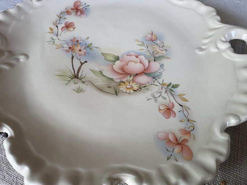 May include: A white ceramic serving platter with a scalloped edge and handles. The platter is decorated with a floral design featuring pink and peach flowers, green leaves, and yellow accents. A decorative piece for serving food.