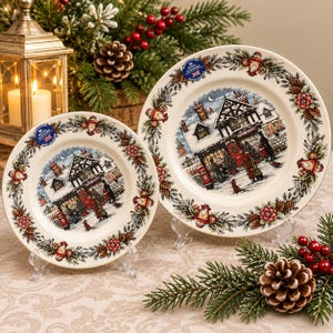 May include: Two cream-colored ceramic plates featuring a central illustration of a snow-covered English village scene with people, a red post box, and a timber-framed building, surrounded by a border of pine cones, holly, and lanterns.