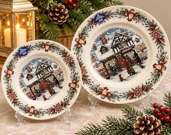 May include: Two cream-colored ceramic plates featuring a central illustration of a snow-covered English village scene with people, a red post box, and a timber-framed building, surrounded by a border of pine cones, holly, and lanterns.