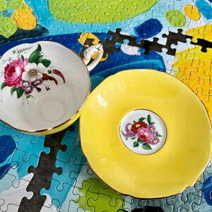 Pu&ograve; includere: Un set di tazza da t&egrave; e piattino in porcellana con esterno giallo brillante e bordi smerlati rifiniti in oro. L'interno della tazza e il centro del piattino sono decorati con un motivo floreale dettagliato di rose rosa e fiori bianchi.