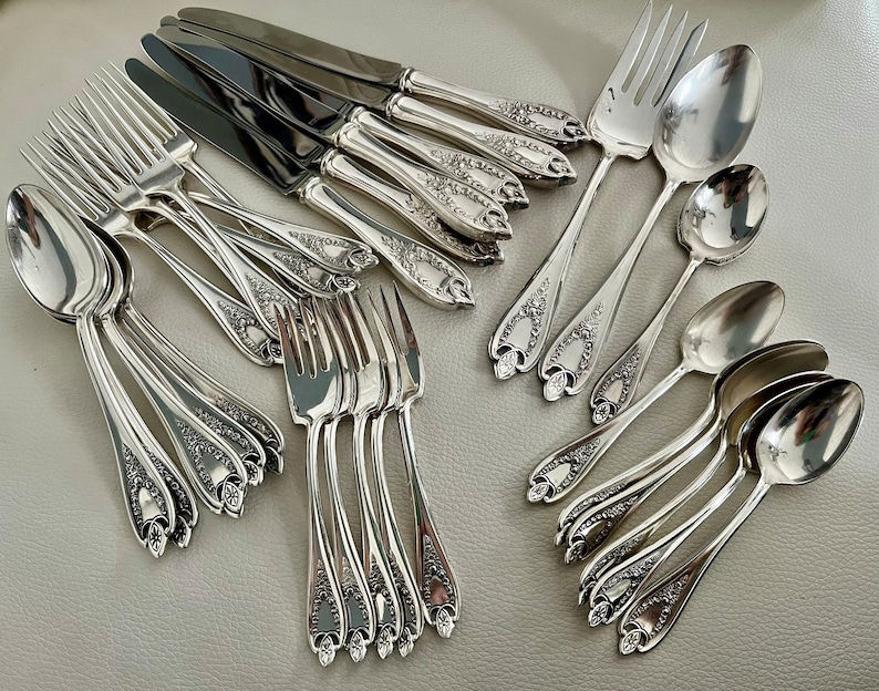 Puede incluir: Un conjunto de cubiertos de plata, que incluye tenedores, cucharas y cuchillos. La cuberter&iacute;a presenta patrones florales decorativos en los mangos. Los cuchillos tienen hojas oscuras y las cucharas y tenedores tienen una superficie brillante y reflectante.