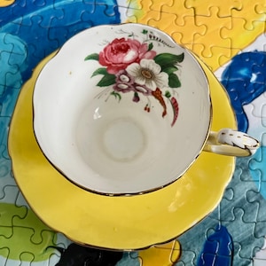 Pu&ograve; includere: Un set composto da tazza da t&egrave; e piattino in porcellana bianca con bordo smerlato e finiture dorate. L'interno della tazza presenta un motivo floreale con fiori rosa e bianchi e foglie verdi. Il piattino &egrave; di un colore giallo brillante.