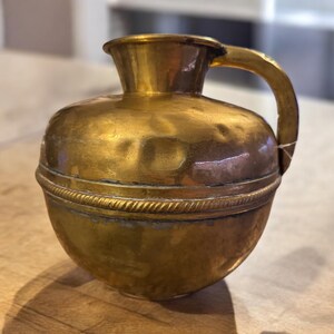 K&ouml;nnte beinhalten: Eine kleine, goldene Messingkanne mit rundem K&ouml;rper und einem einzigen Henkel. Die Kanne hat ein dekoratives Band um die Mitte.