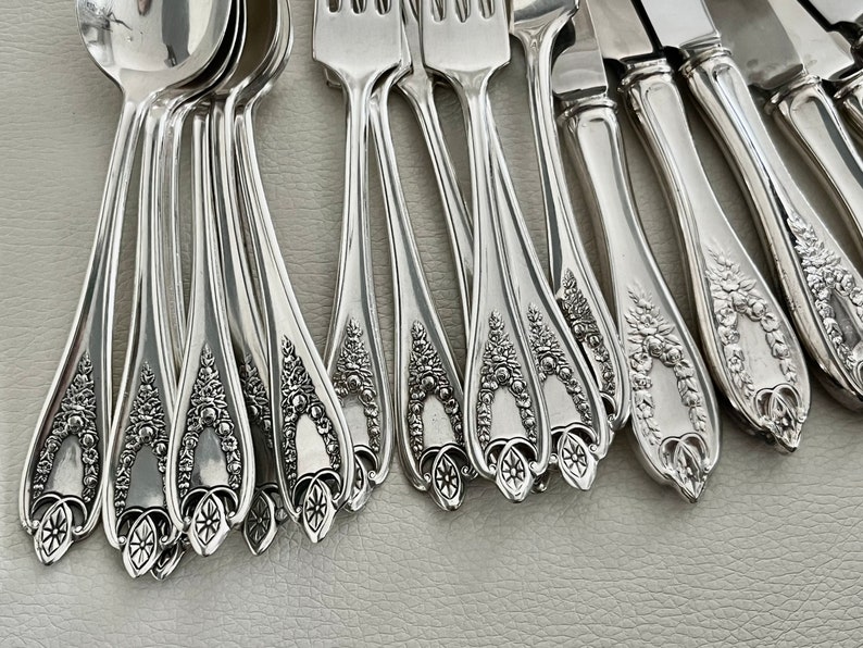 Puede incluir: Una colecci&oacute;n de cubiertos plateados ornamentados, que incluye cucharas, tenedores y cuchillos. Los mangos presentan dise&ntilde;os detallados de flores y volutas. La cuberter&iacute;a est&aacute; dispuesta sobre una superficie gris claro, mostrando la calidad reflectante del metal.