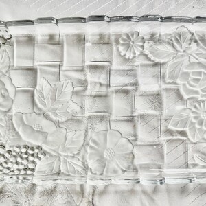 May include: Rectangular clear glass serving tray with a woven basket design. The tray features embossed floral and grape cluster patterns on the edges. The clear glass allows for a view of the contents.