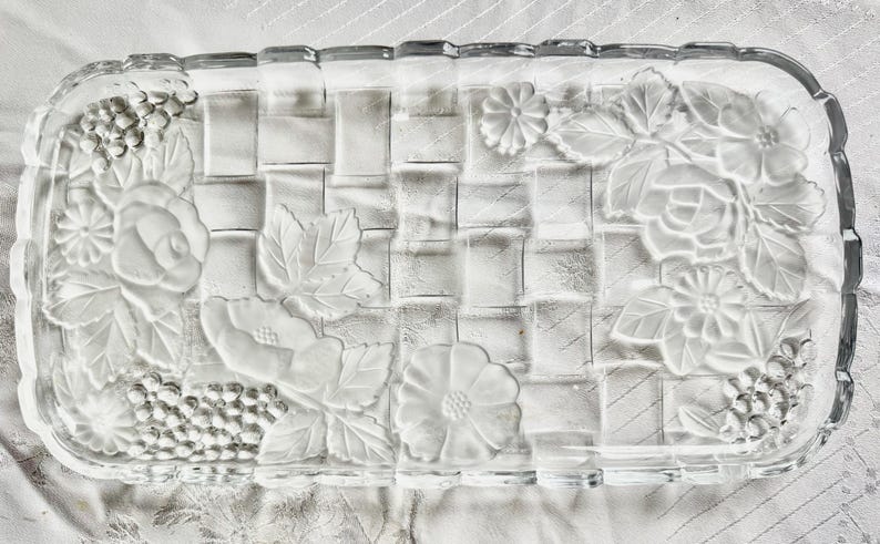 May include: Rectangular clear glass serving tray with a woven basket design. The tray features embossed floral and grape cluster patterns on the edges. The clear glass allows for a view of the contents.